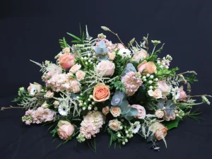Rouwstuk Peach Flowers, een rouw bloemstuk met verschillende bloemen in pastelkleuren zoals zachtroze en lichtblauw, afgewerkt met groen. Dit is een langwerpig en hoog rouwstuk, gemaakt door Bloeibloemen.nl