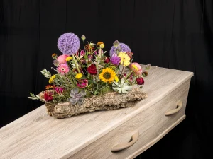 Rouwstuk Robust colour, een rouw bloemstuk op een kist met bloemen in de kleuren paars, geel, rood en roze op een boomschors, gemaakt door Bloeibloemen.nl