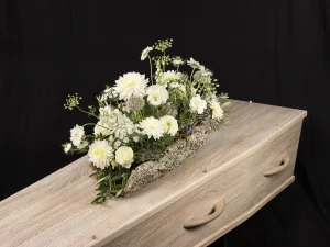 Rouwstuk Robust White, een rouw bloemstuk op een kist met bloemen in witte kleuren op een boomschors, gemaakt door Bloeibloemen.nl