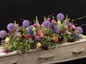 Rouwstuk Sfeer kleuren, een rouw bloemstuk op een kist met bloemen in de kleuren paars, roze, geel, rood, blauw en groen, gemaakt door Bloeibloemen.nl