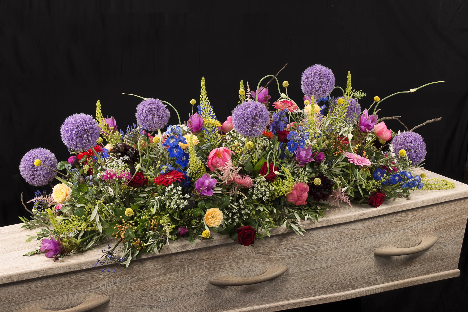 Rouwstuk Sfeer kleuren, een rouw bloemstuk op een kist met bloemen in de kleuren paars, roze, geel, rood, blauw en groen, gemaakt door Bloeibloemen.nl