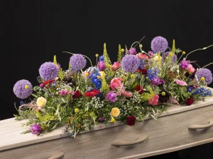 Een kleurrijk afscheid, Sfeer kleuren, een rouw bloemstuk op een kist met bloemen in de kleuren paars, roze, geel, rood, blauw en groen, gemaakt door Bloeibloemen.nl